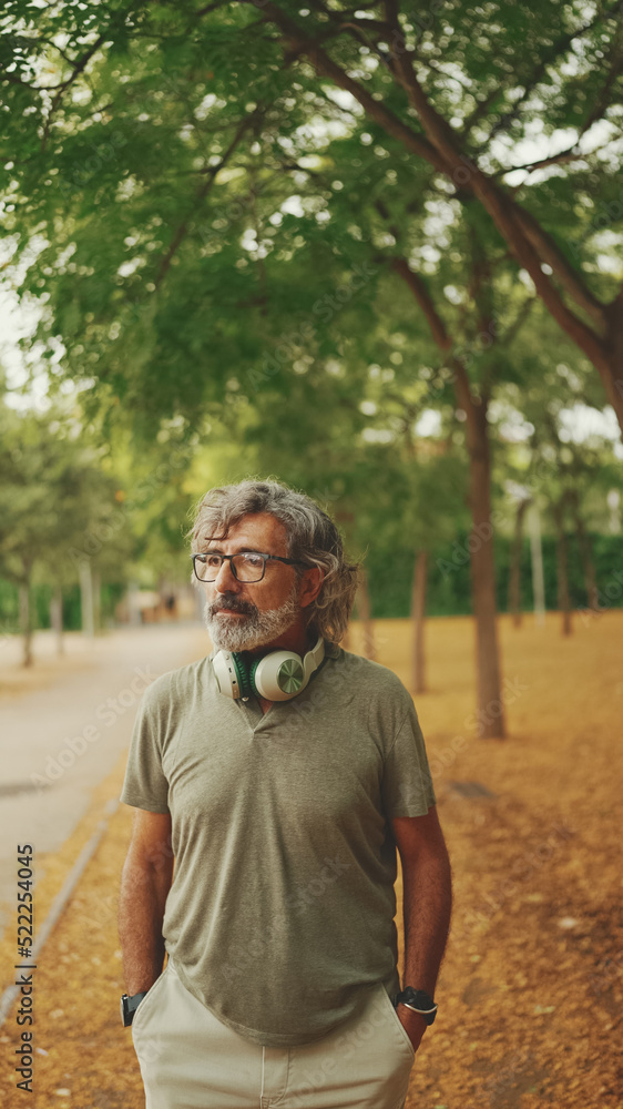 Fototapeta premium Thoughtful middle-aged man with gray hair and beard wearing casual clothes with headphones around his neck is walking down the street