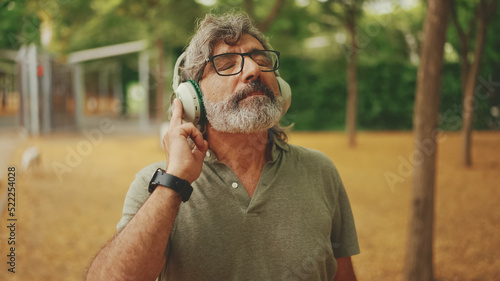 Friendly middle-aged man with gray hair and beard wearing casual clothes listening to music on headphones. Mature gentleman in eyeglasses enjoys music outdoors