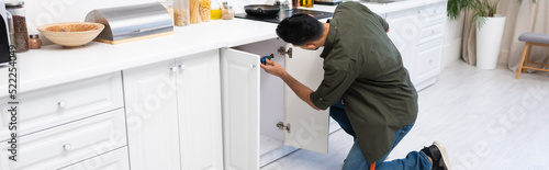 Young muslim man with screwdriver fixing cabinet under worktop in kitchen, banner photo