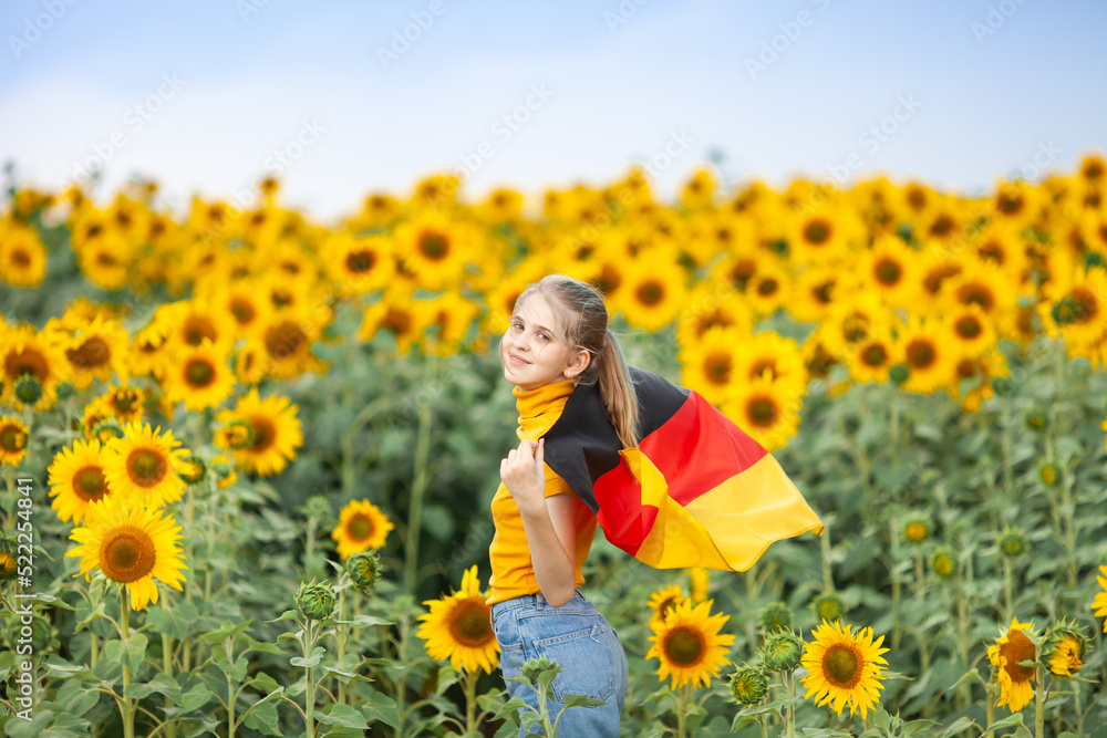 Blond girl holding waving flag of Germany in the sunflower field. Day of German Unity. Travel and learn german language concept. Selective focus. Blond girl holding waving flag of Germany in the sunflower field. Day of German Unity. Travel and learn german language concept. Selective focus.