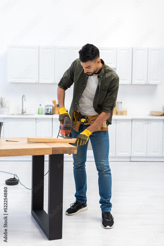 Young muslim craftsman holding jigsaw machine near wooden plank in kitchen Young muslim craftsman holding jigsaw machine near wooden plank in kitchen