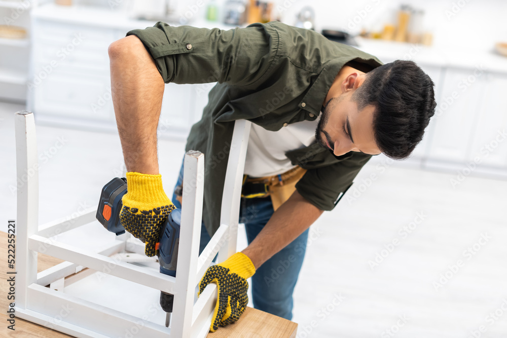 Muslim man in gloves using electric screwdriver while fixing chair in kitchen Muslim man in gloves using electric screwdriver while fixing chair in kitchen