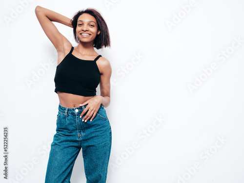 Portrait of young beautiful black woman. Smiling model dressed in summer jeans clothes. Sexy carefree female posing near white wall in studio. Tanned and cheerful