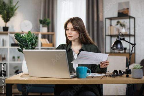Likable young Caucasian woman sitting at desk with laptop and holding papers. Pleasant female in casual shirt using portable computer for work at bright room at home.