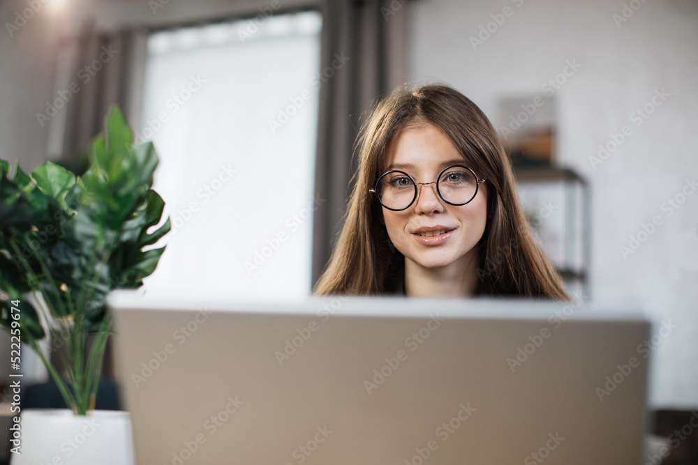 Beautiful caucasian woman in glasses with long dark hair working on wireless laptop while sitting at desk. Female freelancer in casual shirt using computer for remote work. Beautiful caucasian woman in glasses with long dark hair working on wireless laptop while sitting at desk. Female freelancer in casual shirt using computer for remote work.
