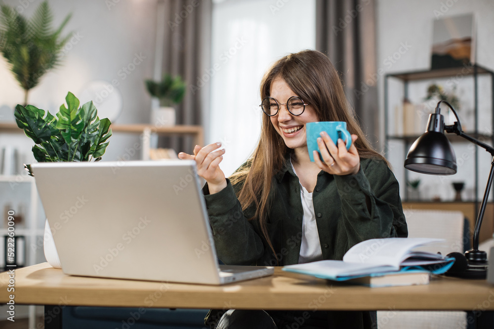 Excited young caucasian woman holding cup with hot drink and laptop for video conversation at office. Female freelancer sitting at table and taking notes during video chat. Excited young caucasian woman holding cup with hot drink and laptop for video conversation at office. Female freelancer sitting at table and taking notes during video chat.