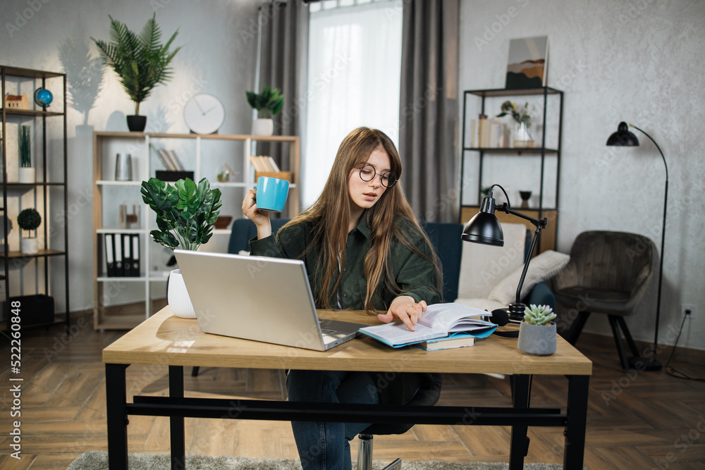 Smiling Caucasian woman in eyeglasses and denim shirt, posing indoors at bright office or home interior. Pretty lady with hot drink reading notes while working at laptop. Smiling Caucasian woman in eyeglasses and denim shirt, posing indoors at bright office or home interior. Pretty lady with hot drink reading notes while working at laptop.