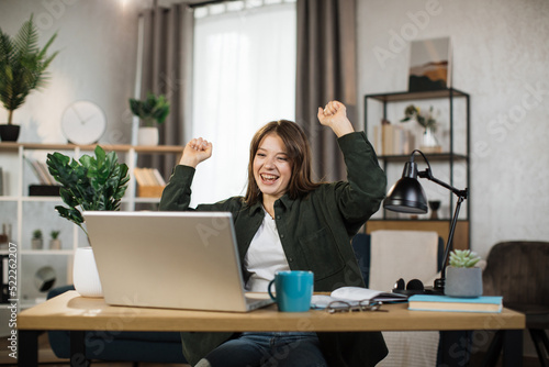 Happy caucasian woman student in casual wear sitting at table with modern laptop and keeping arms raised. Concept of people, technology and distance learning and success.