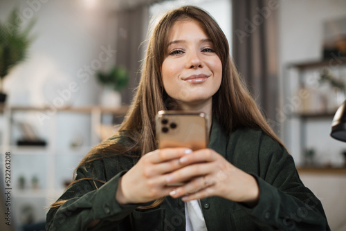 Smiling businesswoman using modern smartphone for video chat at work. Young caucasian female sitting at modern bright office. Technology concept.