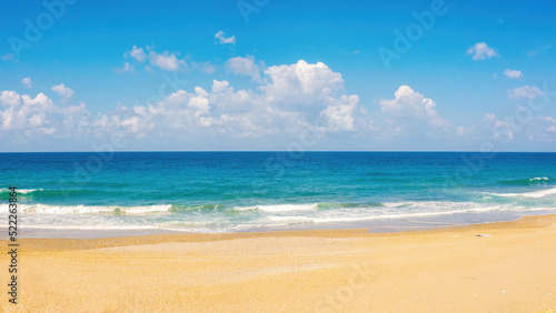 Beautiful background widescreen image of the surf with small caps of white foam, yellowish beach sand and blue sky with clouds. © Laura Pashkevich