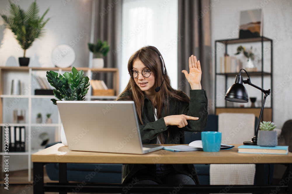 Young caucasian woman using headset and laptop for video conversation at office. Female freelancer sitting at table and talking with client during video chat. Young caucasian woman using headset and laptop for video conversation at office. Female freelancer sitting at table and talking with client during video chat.