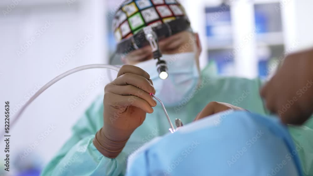 Male surgeon in mask, cap and flashlight on his head operates the patient. Doctor applies otolaryngology instrument to a patient’s nose. Male surgeon in mask, cap and flashlight on his head operates the patient. Doctor applies otolaryngology instrument to a patient’s nose.
