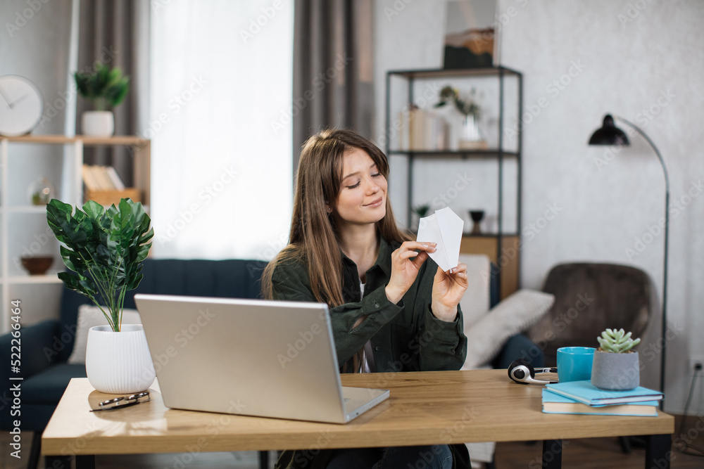Attractive caucasian woman in casual outfit sitting at desk with modern laptop and playing with paper plane. Female boring at work. Unproductive concept. Attractive caucasian woman in casual outfit sitting at desk with modern laptop and playing with paper plane. Female boring at work. Unproductive concept.
