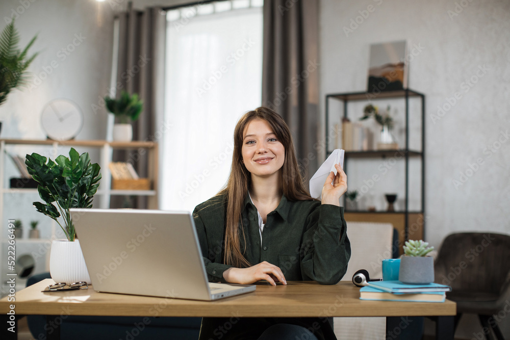 Smiling female freelancer playing with paper plane while working from home. Attractive caucasian woman taking break during working time.
