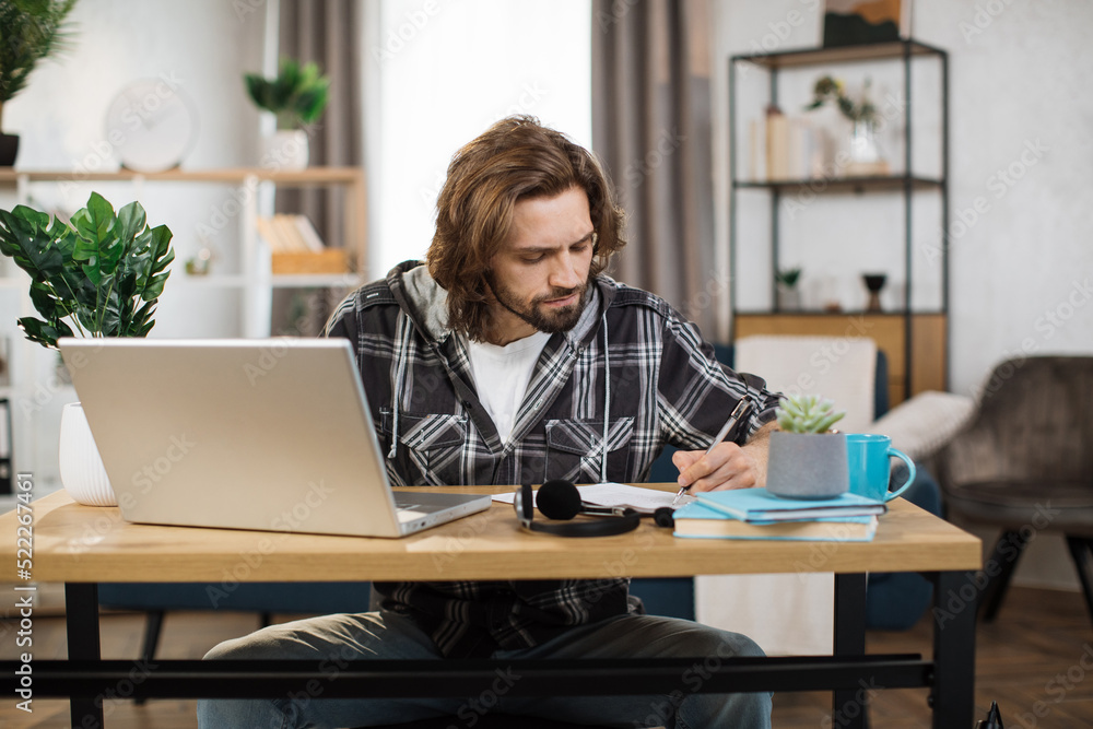 Focused bearded male student in casual wear sitting at table with modern laptop writing notes. Concept of people, technology and distance learning. Focused bearded male student in casual wear sitting at table with modern laptop writing notes. Concept of people, technology and distance learning.