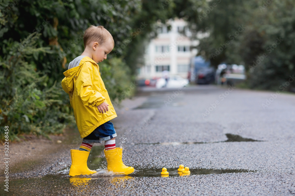 A small child in rainbow socks, yellow rubber boots and a jacket jumps through puddles and plays with yellow rubber ducks. A picture of summer and autumn holidays. A child in the rain.