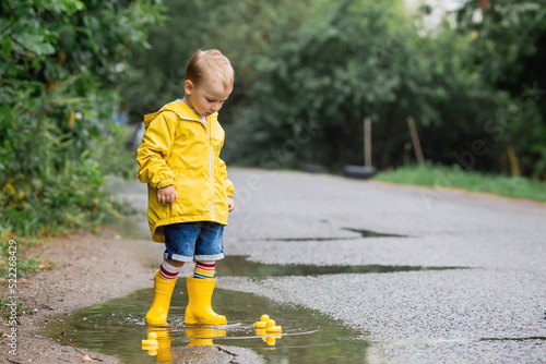 A small child in rainbow socks, yellow rubber boots and a jacket jumps through puddles and plays with yellow rubber ducks. A picture of summer and autumn holidays. A child in the rain.