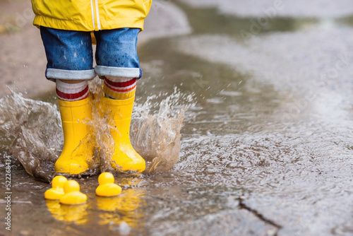 A small child in rainbow socks, yellow rubber boots and a jacket runs through puddles, has fun and plays after the rain. A picture of summer and autumn holidays. Legs close-up.