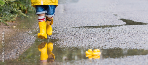 A small child in rainbow socks, yellow rubber boots and a jacket runs through puddles, has fun and plays after the rain. A picture of summer and autumn holidays. Legs close-up. photo