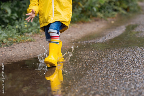 A small child in rainbow socks, yellow rubber boots and a jacket runs through puddles, has fun and plays after the rain. A picture of summer and autumn holidays. Legs close-up.