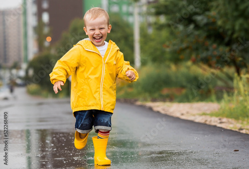 A small child in rainbow socks, yellow rubber boots and a jacket runs through puddles, has fun and plays after the rain. A picture of summer and autumn holidays. A happy child.