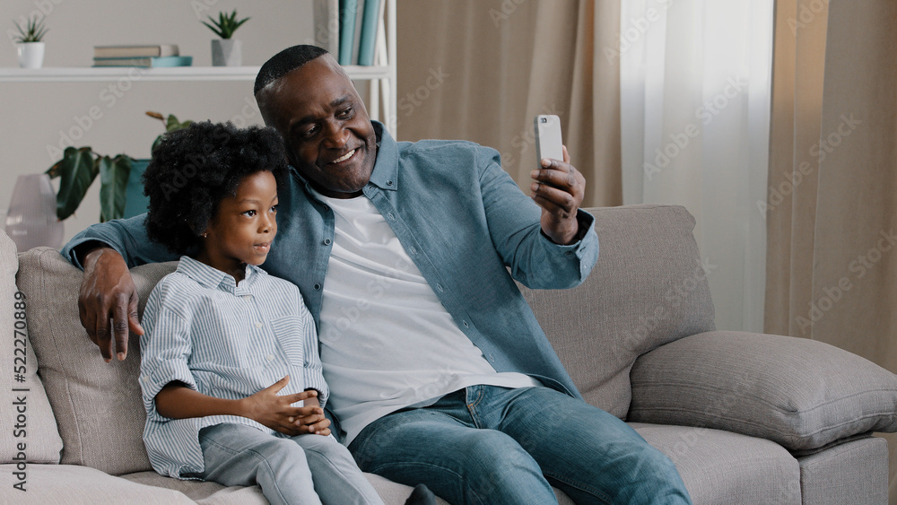 African American family father and daughter sitting in room on couch chatting on webcam mature man with little cute girl shoots video for blog looking at smartphone screen waving hello to phone camera African American family father and daughter sitting in room on couch chatting on webcam mature man with little cute girl shoots video for blog looking at smartphone screen waving hello to phone camera