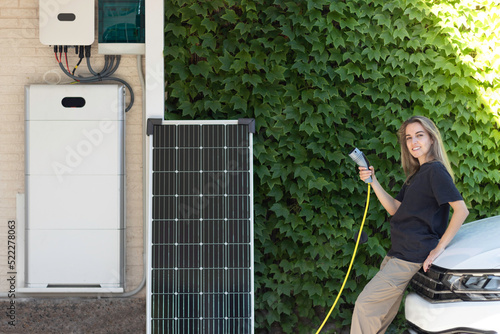 Young beautiful woman looking to camera and smiling on white electric car holding a plug and a solar system on the side