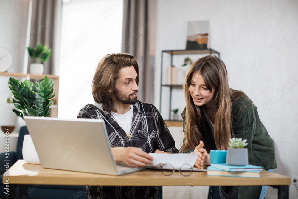 Beautiful caucasian woman standing near bearded man that sitting at table and using laptop and showing information on papers. Two office workers in casual outfit. Work with modern technology. Beautiful caucasian woman standing near bearded man that sitting at table and using laptop and showing information on papers. Two office workers in casual outfit. Work with modern technology.
