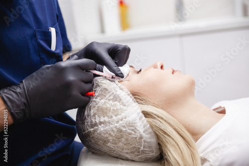 A cosmetologist in black gloves injects botulinum toxin into the patient's forehead. A blonde patient in a disposable cap lies with her eyes closed