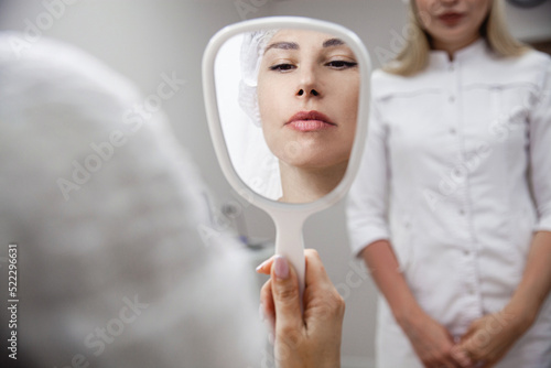 A young woman of European appearance looks in the mirror after the rejuvenating procedures in the beauty salon. In the background is a cosmetologist in a white coat photo