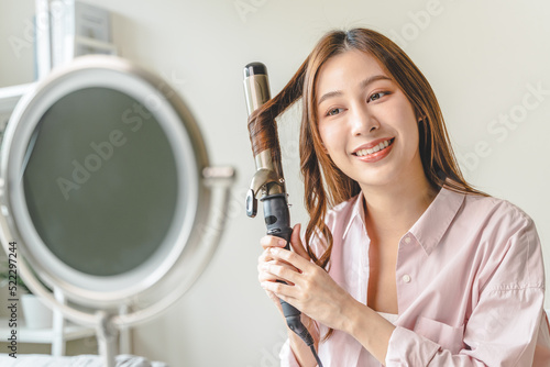 Fototapeta women hairdo makeup routine, Young woman looking at the mirror and using iron hair curling curly long hair at home