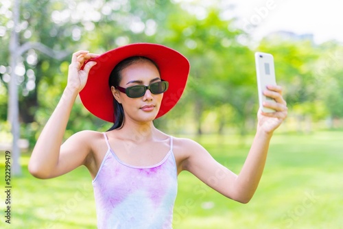 Portrait photo of the moment of a young asian beautiful lady with a red hat and sunglasses happily doing video call on her smartphone with her friends during a garden park strolling