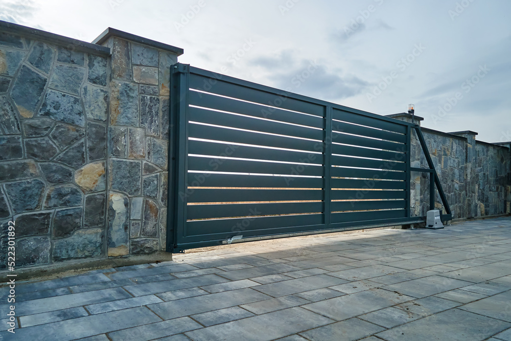 Wide automatic sliding gate with remote control installed in high stone fense wall. Security and protection concept Wide automatic sliding gate with remote control installed in high stone fense wall. Security and protection concept