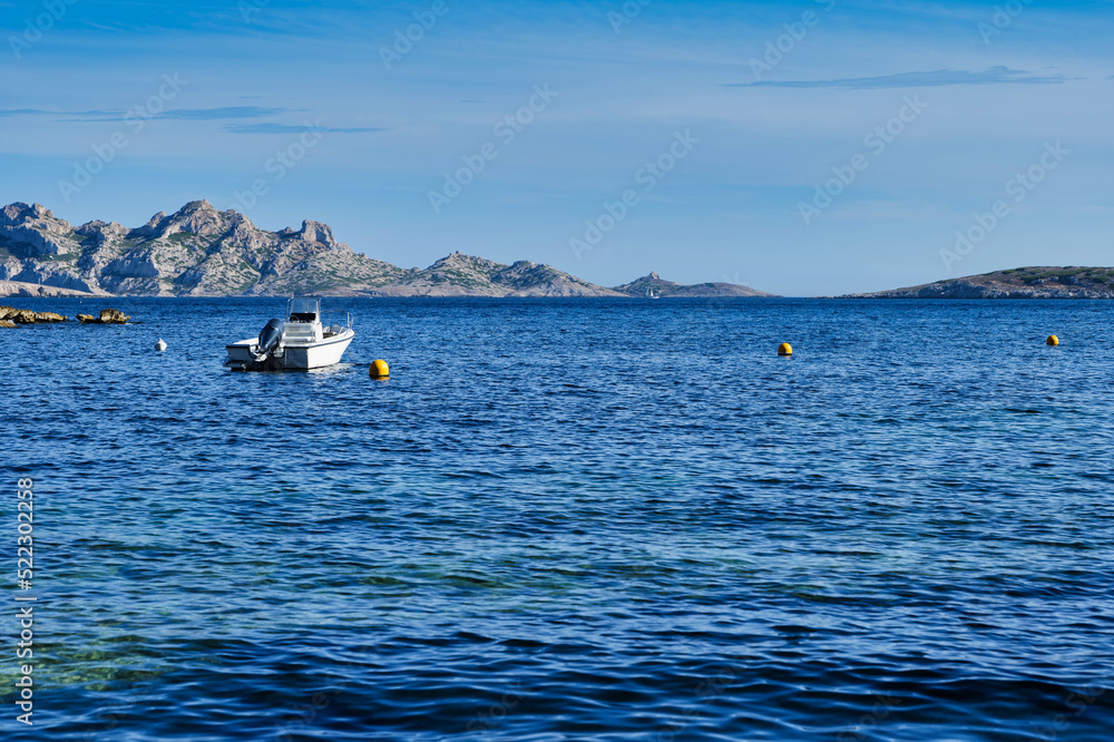 Small recreational boat tied to a yellow buoy in a bay close to Marseille, France. Small rocky islands in the background. Small boat on the dark blue ocean. 
