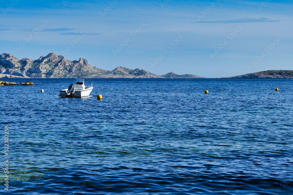 Obraz premium Small recreational boat tied to a yellow buoy in a bay close to Marseille, France. Small rocky islands in the background. Small boat on the dark blue ocean. 