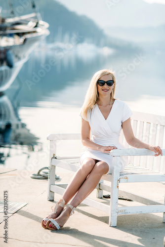 young sexy smiling caucasian woman with sunglasses sits on sea shore in white dress.