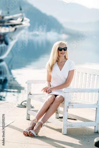 young sexy smiling caucasian woman with sunglasses sits on sea shore in white dress.