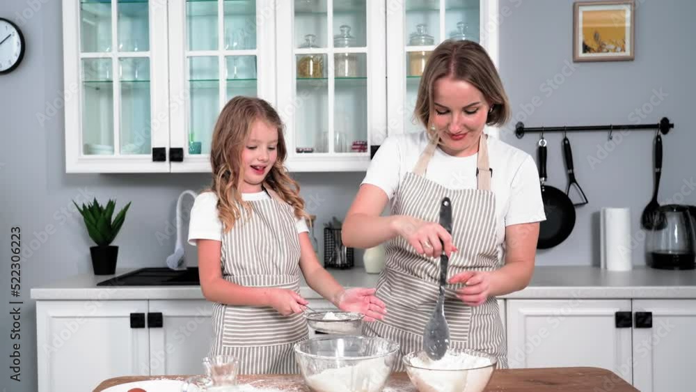 portrait of charming girl together with her mother pour wheat flour into bowl for making bun, smile and look at camera while standing at table backdrop of kitchen portrait of charming girl together with her mother pour wheat flour into bowl for making bun, smile and look at camera while standing at table backdrop of kitchen