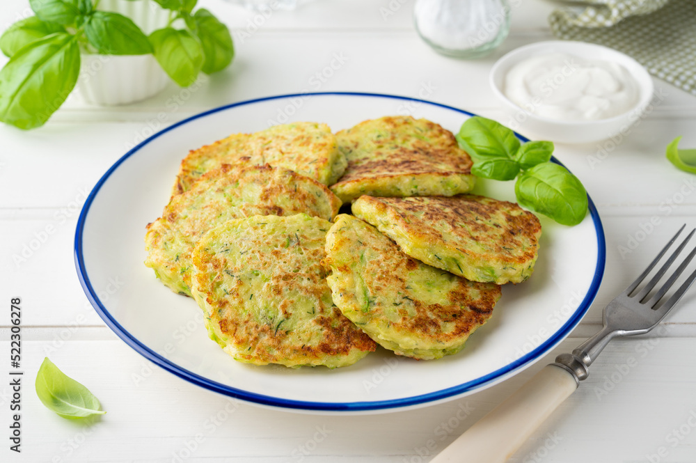 Zucchini fritters with cheese, garlic and herbs. Vegetarian zucchini pancakes, served with basil and sour cream on a white wooden background. Selective focus. Zucchini fritters with cheese, garlic and herbs. Vegetarian zucchini pancakes, served with basil and sour cream on a white wooden background. Selective focus.