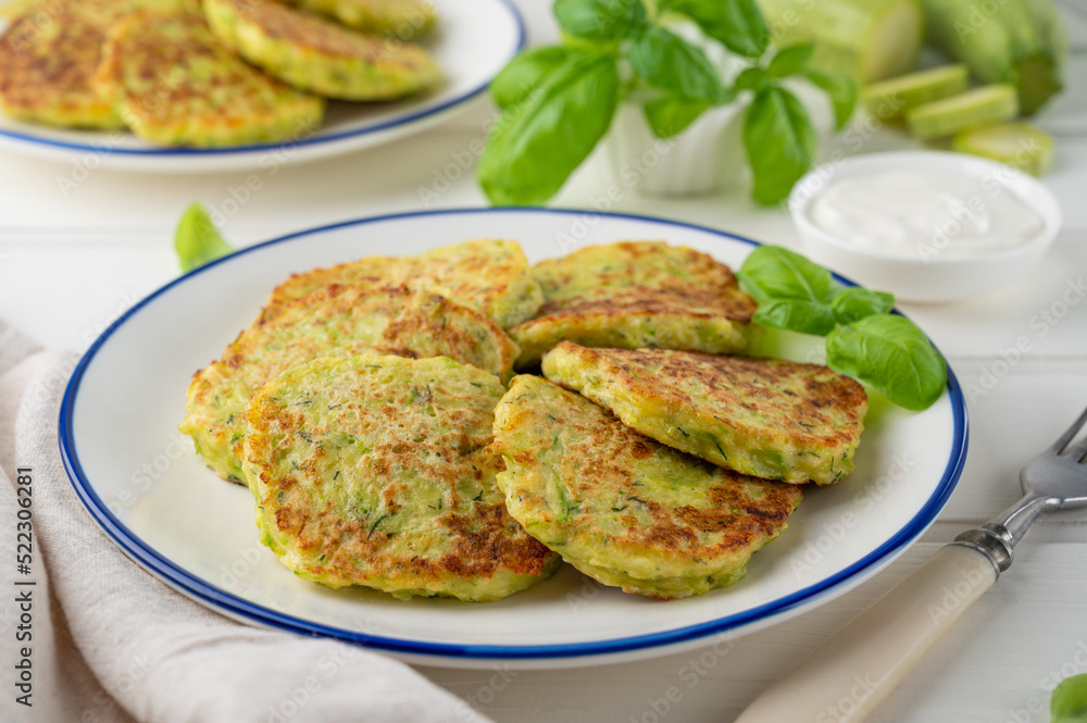 Zucchini fritters with cheese, garlic and herbs. Vegetarian zucchini pancakes, served with basil and sour cream on a white wooden background. Selective focus.