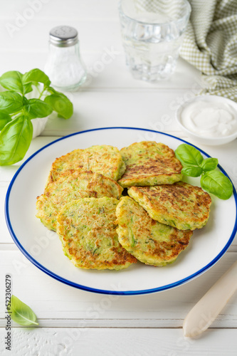 Zucchini fritters with cheese, garlic and herbs. Vegetarian zucchini pancakes, served with basil and sour cream on a white wooden background. Selective focus.