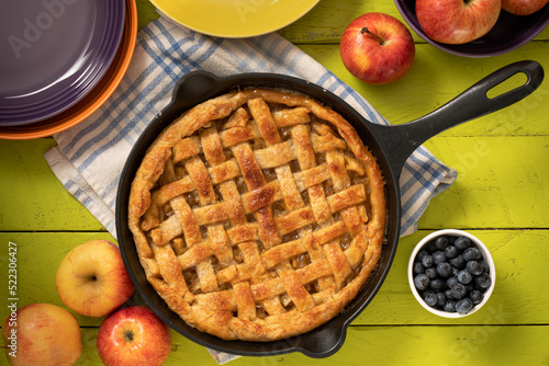 Apple pie in a cast iron skillet on a green coor table surrounded by apples. photo