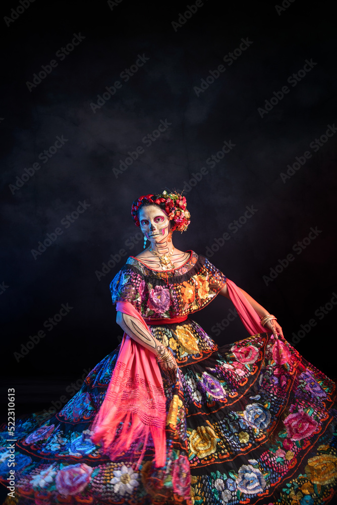 catrina woman dressed in a mexican chiapas costume with a black background pink rebozo and skull and bones makeup on her hands