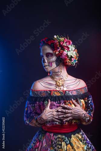 catrina woman dressed in a mexican chiapas costume with a black background pink rebozo and skull and bones makeup on her hands