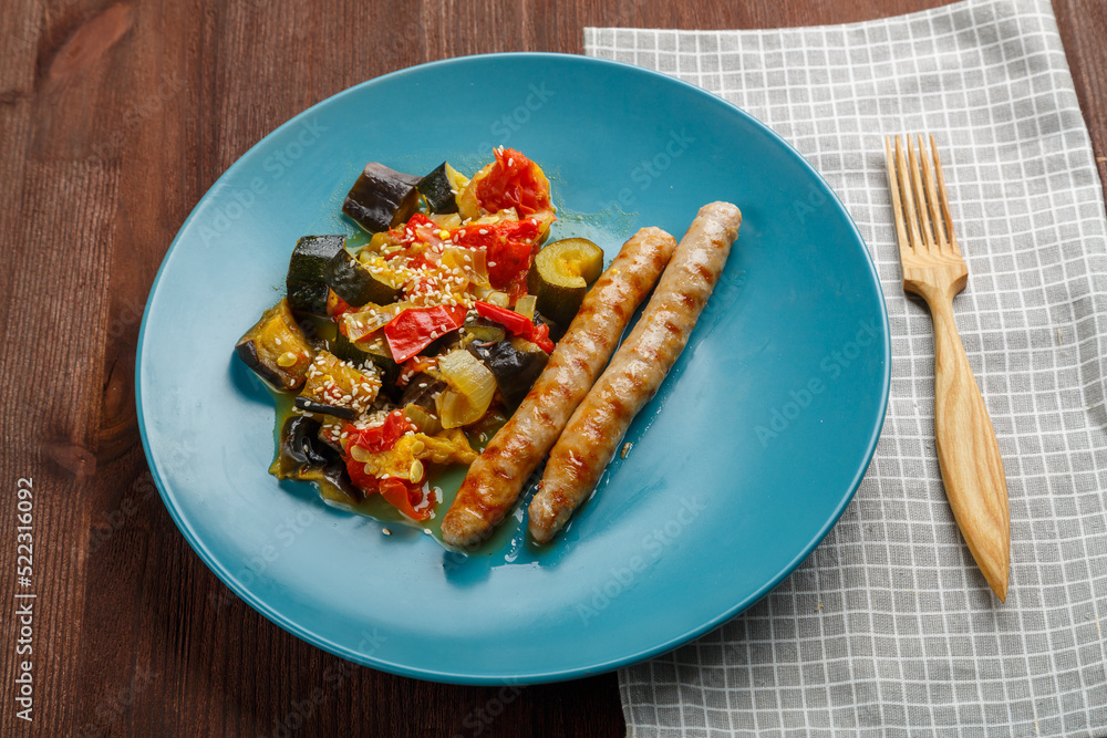 A plate of vegetables with grilled sausages on a table on a napkin and a wooden fork.