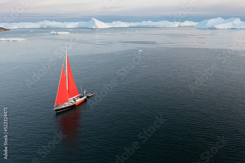 Sail boat with red sails cruising among ice bergs during dusk in front of a full moon. Disko Bay, Greenland. Midnight sun, romantic view. Climate change and global warming © Michal
