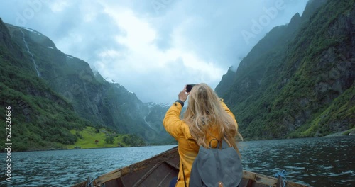 Female travel researcher taking phone photos of green mountain views, sitting alone in a row boat. Back view of woman doing ecology research outside. Traveler takes a picture for online content