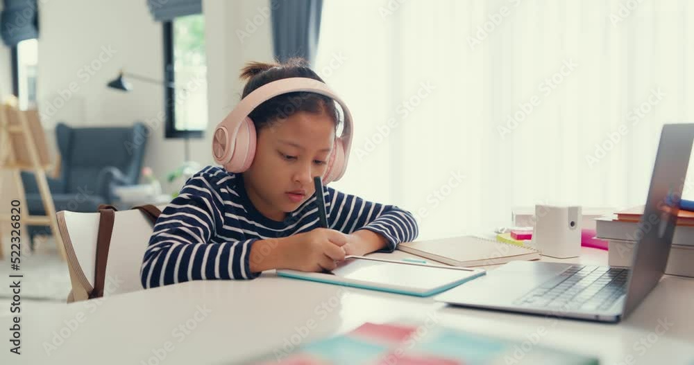 Asian toddler girl with sweater wear headphone sit front of desk with notepad use magic pen focus on writing do homework, finger counting number math online learning course from laptop at home. Asian toddler girl with sweater wear headphone sit front of desk with notepad use magic pen focus on writing do homework, finger counting number math online learning course from laptop at home.