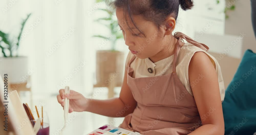 Close-up Asian toddler girl with apron sit on sofa chair in front of painting canvas with color palette use paint brush focus on painting abstract on weekend at home. Creative lifestyle kid concept. Close-up Asian toddler girl with apron sit on sofa chair in front of painting canvas with color palette use paint brush focus on painting abstract on weekend at home. Creative lifestyle kid concept.