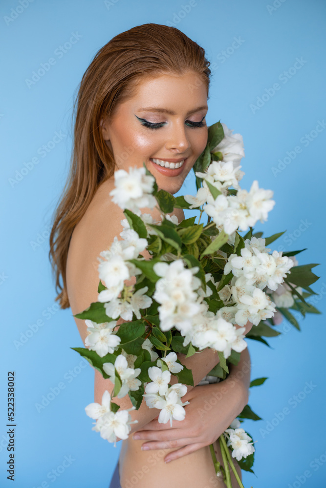 Portrait of young smiling naked stunning ginger woman standing on blue background, holding white flowers of mock orange. Portrait of young smiling naked stunning ginger woman standing on blue background, holding white flowers of mock orange.